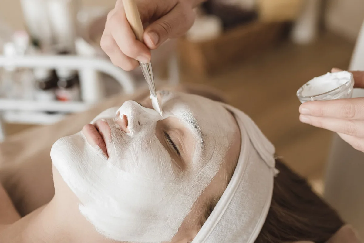 An esthetician brushes a thick white mask onto a client's face while holding a small glass bowl