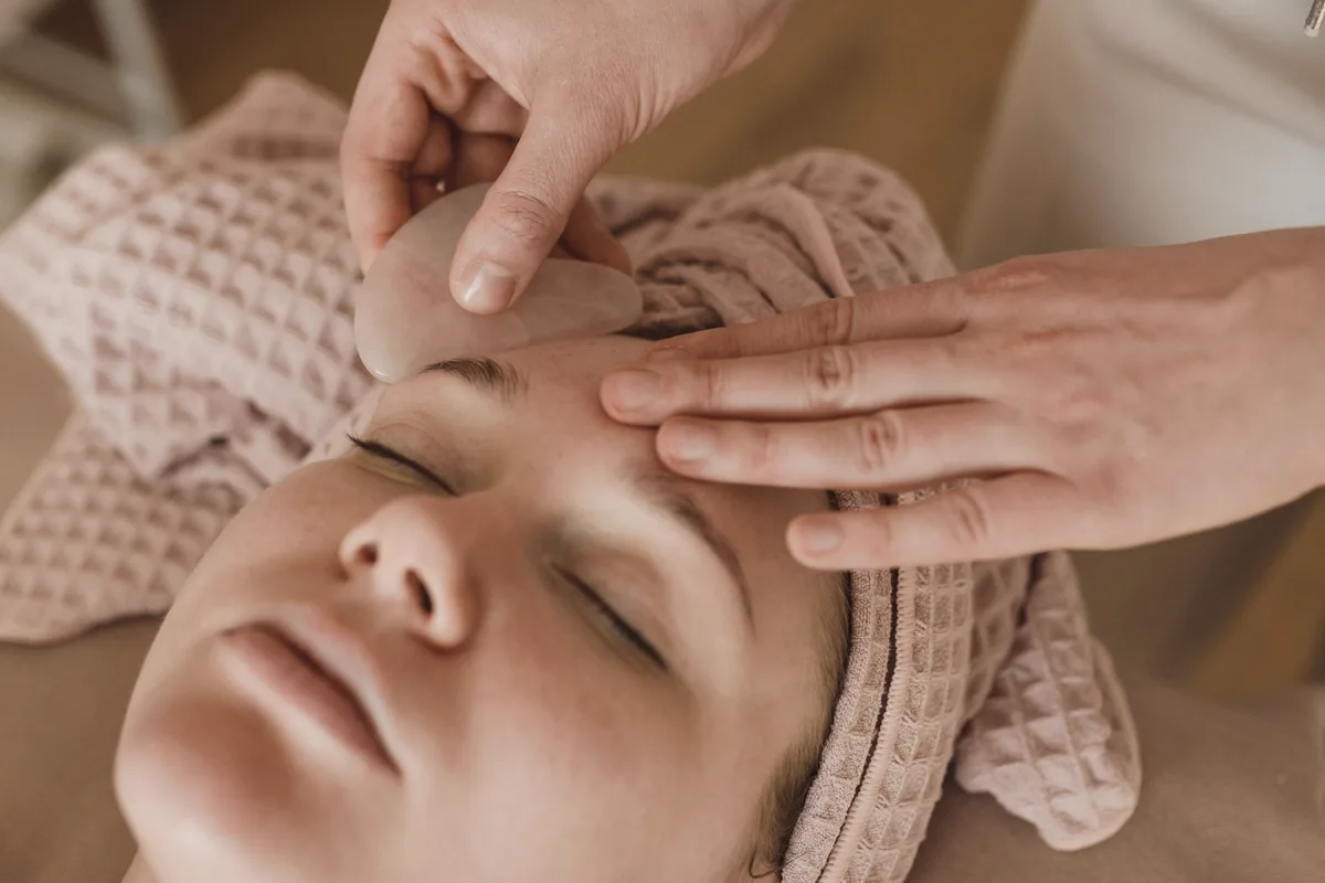 An esthetician's hands gliding a pink facial stone across a relaxed client's forehead